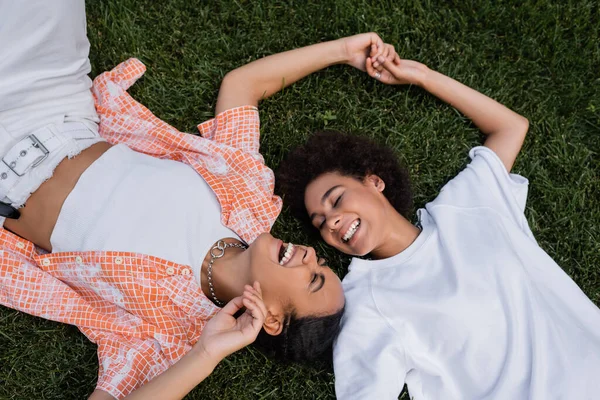Vista superior de las mujeres lesbianas afroamericanas felices cogidas de la mano y tumbadas en la hierba - foto de stock