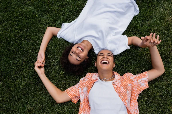 Vista superior de la pareja de lesbianas afroamericanas positivas cogidas de la mano y tumbadas en la hierba - foto de stock