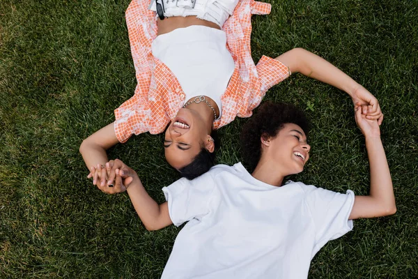 Vista superior de las sonrientes mujeres lesbianas afroamericanas cogidas de la mano y tumbadas en la hierba - foto de stock