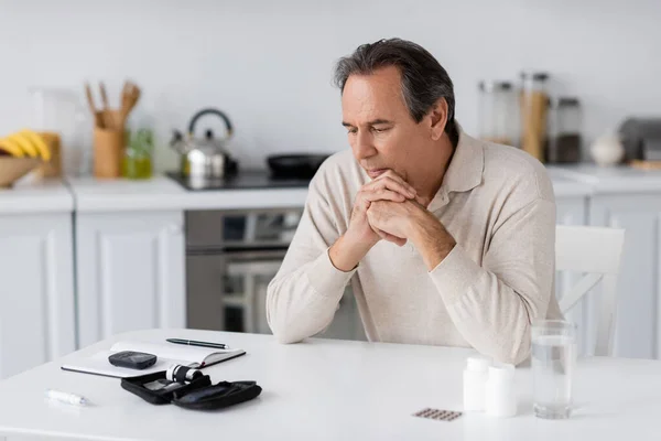 Nachdenklicher und zuckerkranker Mann mittleren Alters blickt auf Glukosemessgerät und Lanzettenstift auf dem Tisch — Stockfoto