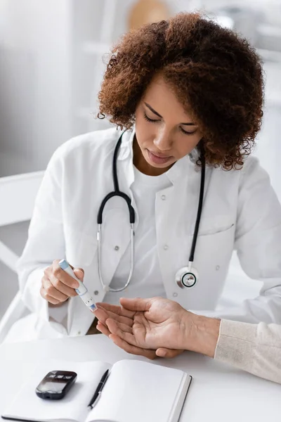 Кудрявый африканский американский врач берёт образец крови взрослого пациента с ручкой Ланцета возле глюкозы на столе — стоковое фото