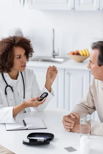 Médecin afro-américain bouclé tenant le dispositif glucomètre et parlant avec un patient d'âge moyen — Photo de stock