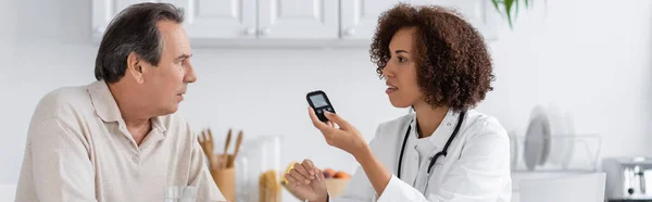 Vue latérale du médecin afro-américain bouclé tenant le dispositif de glucomètre et parlant avec un patient d'âge moyen, bannière — Photo de stock