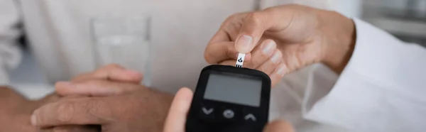 Vue partielle du médecin afro-américain montrant la bande de test sur le dispositif de glucomètre au patient d'âge moyen, bannière — Photo de stock