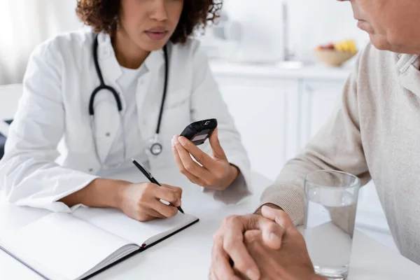 Cropped view of african american doctor holding glucometer and writing in notebook near middle aged patient — Stock Photo