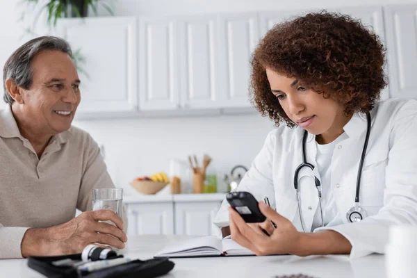 Curly african american doctor holding glucometer and writing in notebook near smiling middle aged patient — Stock Photo