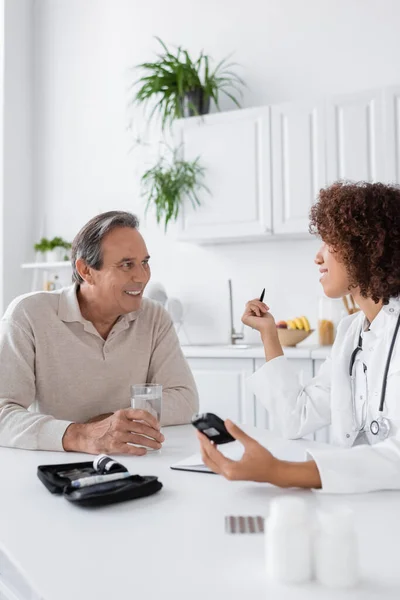 Curly african american doctor holding glucometer near smiling middle aged patient — Stock Photo