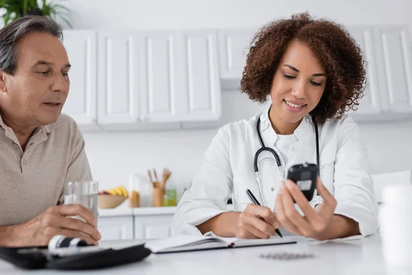 Lächelnder afrikanisch-amerikanischer Arzt mit Glukometer und Notizbuch in der Nähe eines Patienten mittleren Alters — Stockfoto