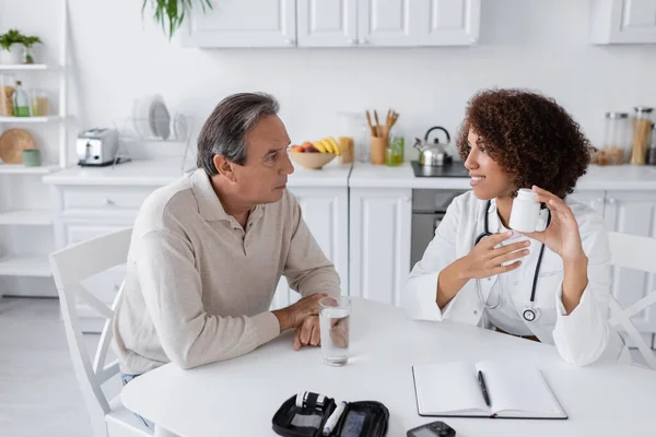 Smiling african american doctor in white coat holding bottle with pills near middle aged patient with diabetes — Stock Photo