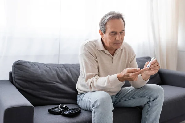 Middle aged man taking blood sample with lancet pen while sitting on couch in living room — Stock Photo