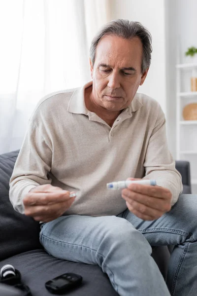 Зрелый мужчина с сахарным диабетом, держащий перо ланцета и тестовую полосу, сидя на диване в гостиной — стоковое фото