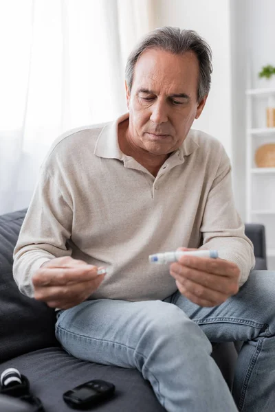 Mature man with diabetes holding lancet pen and test strip while sitting on couch in living room — Stock Photo