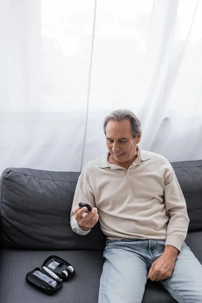 Весёлый мужчина средних лет с сахарным диабетом, держит устройство для измерения уровня глюкозы и сидит на диване — стоковое фото