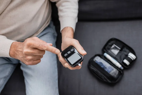 Partial view of middle aged man with diabetes pointing at glucose meter device and sitting on sofa — Stock Photo