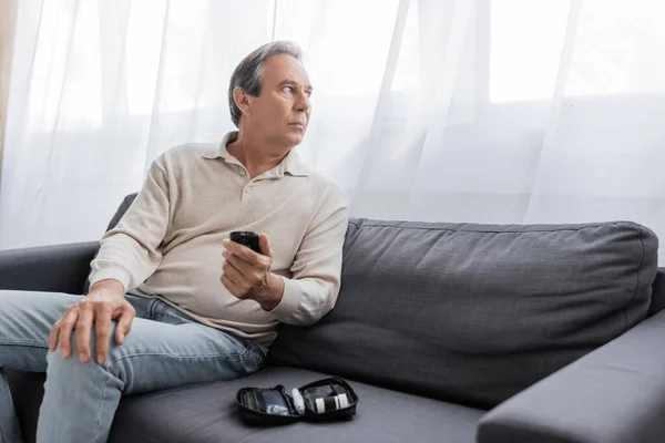 Мужчина среднего возраста с сахарным диабетом держит устройство глюкозы и сидит на диване в гостиной — стоковое фото