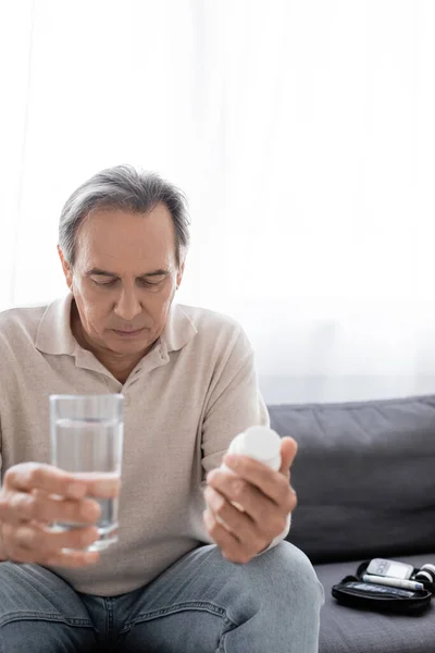 Triste homme d'âge moyen avec le diabète tenant verre d'eau et des pilules tout en étant assis sur le canapé — Photo de stock