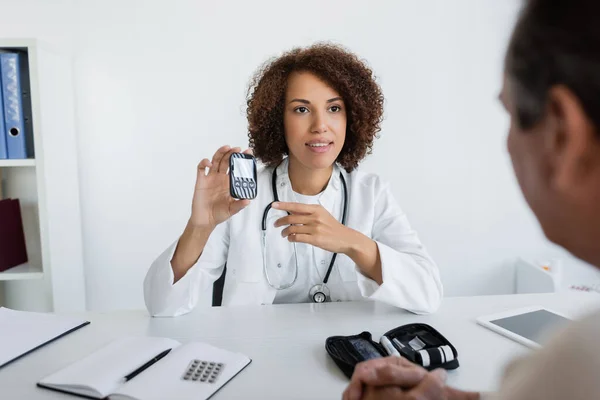 Young african american doctor pointing at glucometer near middle aged patient in clinic — Stock Photo