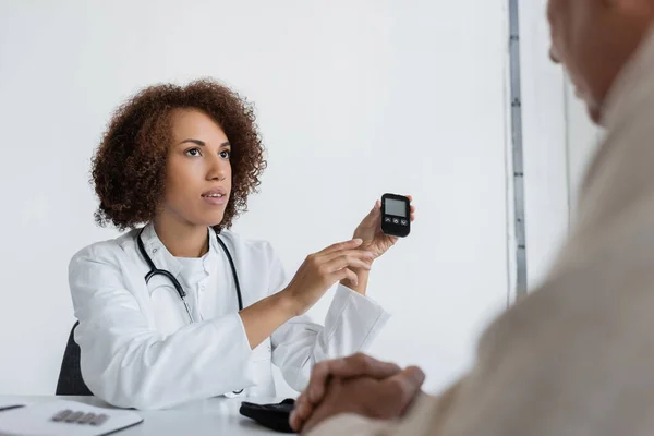Médecin afro-américain bouclé tenant dispositif de glycomètre près de l'homme d'âge moyen avec diabète en clinique — Photo de stock