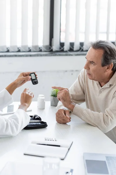 Middle aged man with diabetes pointing with finger at glucose meter device near african american doctor in clinic — Stock Photo