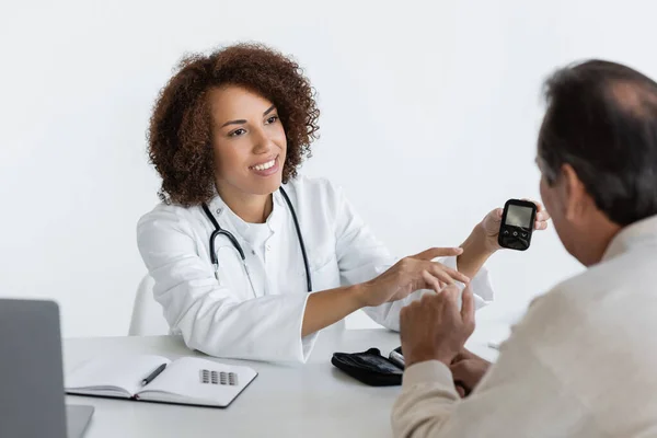 Alegre afroamericano doctor apuntando a glucosímetro cerca de mediana edad hombre con diabetes en clínica - foto de stock