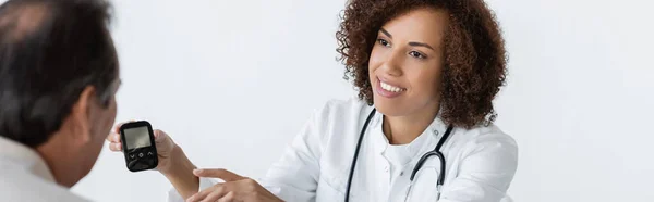 Joyeux médecin afro-américain pointant du doigt le glucomètre près d'un homme d'âge moyen diabétique à la clinique, bannière — Photo de stock