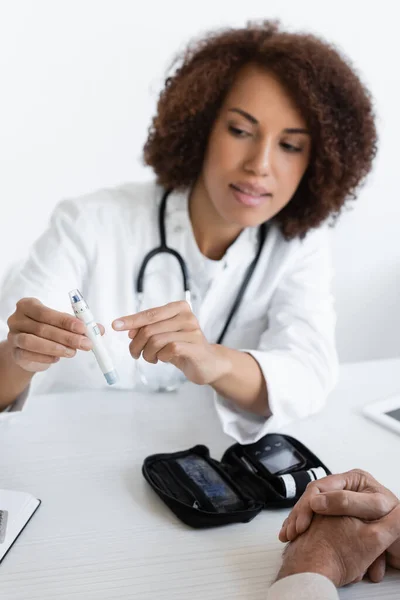 Médico afroamericano señalando con el dedo a lápiz lanceta cerca de paciente de mediana edad con diabetes en la clínica - foto de stock