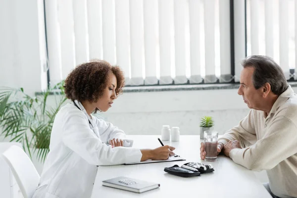 African american doctor writing on clipboard near mature patient with diabetes holding glass of water in clinic — Stock Photo