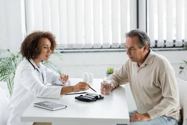 African american doctor pointing at glucometer device with pen near mature patient with diabetes holding glass of water — Stock Photo