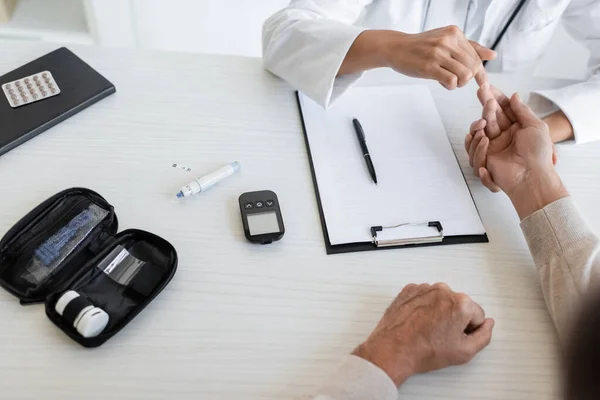 Vista recortada del médico afroamericano revisando la mano del paciente maduro cerca del kit de diabetes - foto de stock