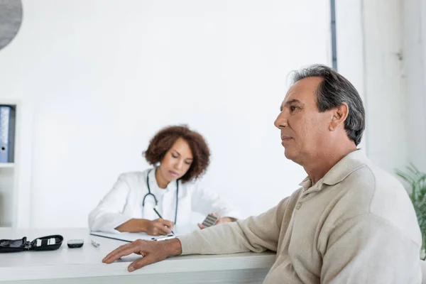 Homme mûr avec diabète assis près flou afro-américain médecin en clinique — Photo de stock