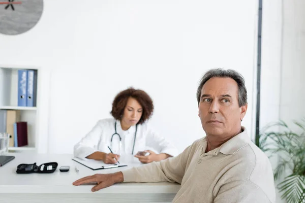 Mature man with diabetes sitting near blurred african american doctor in clinic — Stock Photo