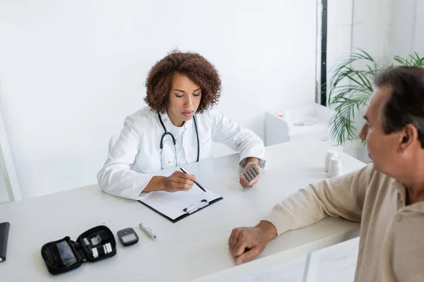Afrikanisch-amerikanischer Arzt hält Tabletten in der Hand und blickt auf Klemmbrett in der Nähe eines Patienten mit Diabetes in der Klinik — Stockfoto