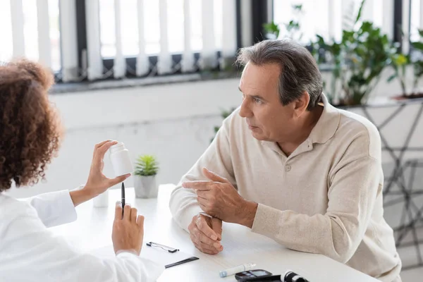 Mature patient with diabetes pointing at pills near african american doctor in clinic — Stock Photo