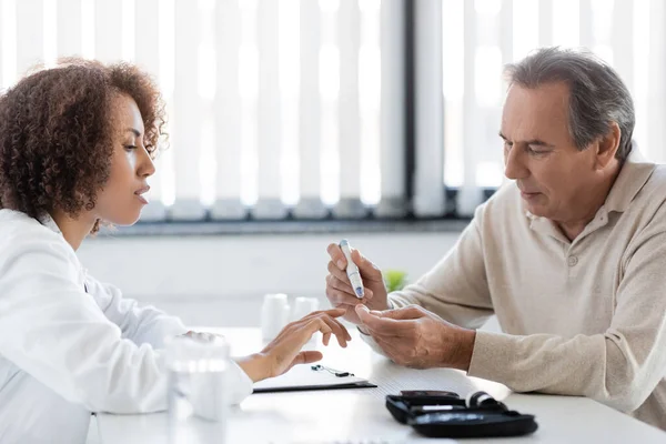 African american doctor looking at mature patient using lancet pen in clinic — Stock Photo
