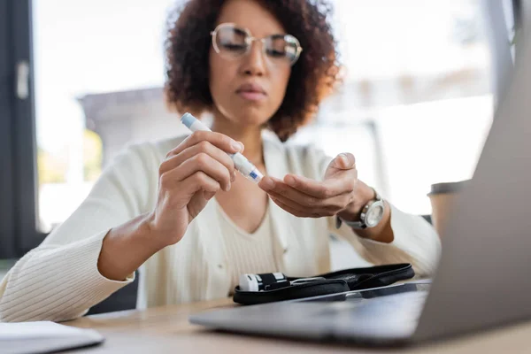 Femme d'affaires afro-américaine floue utilisant un stylo lancette près d'un ordinateur portable et un kit diabète au bureau — Photo de stock