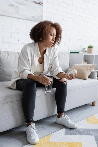 Femme afro-américaine diabétique regardant des pilules tout en tenant un verre d'eau à la maison — Photo de stock