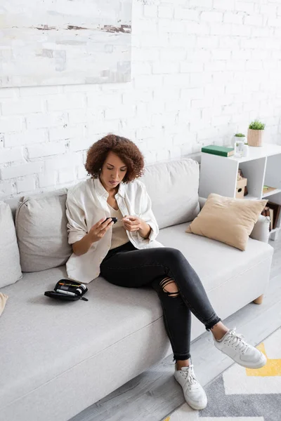 Young african american woman with diabetes using glucometer on couch at home — Stock Photo