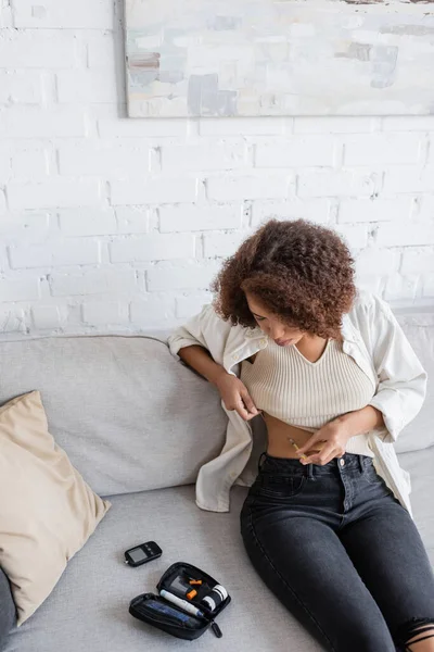 African american woman with diabetes doing insulin injection near medical kit on couch — Stock Photo