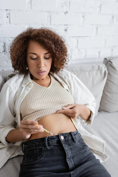 African american woman with diabetes doing insulin injection while sitting on couch in living room — Stock Photo