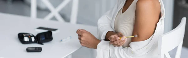Обрезанный вид на африканскую американку, делающую инъекцию инсулина рядом с размытым глюкометром на кухне, баннер — стоковое фото