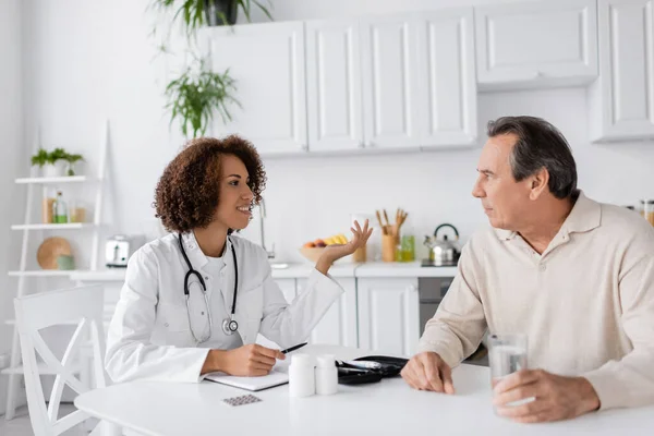 Smiling african american doctor talking to mature patient with diabetes near pills at home — Stock Photo