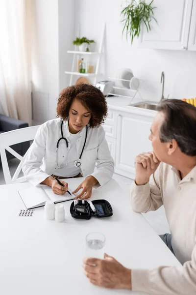 African american doctor writing on notebook near diabetes kit and patient at home — Stock Photo