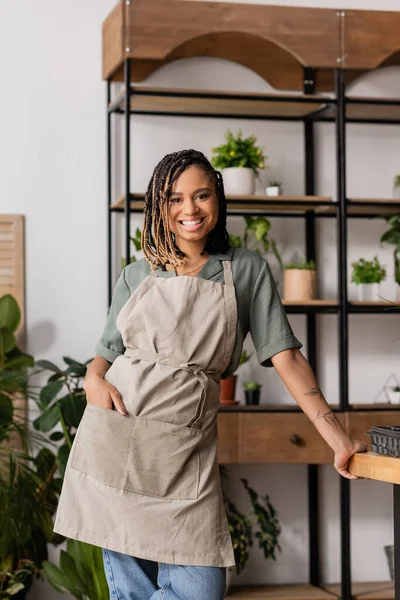 Stylish african american woman holding hand in pocket of apron and smiling at camera in flower shop — Stock Photo
