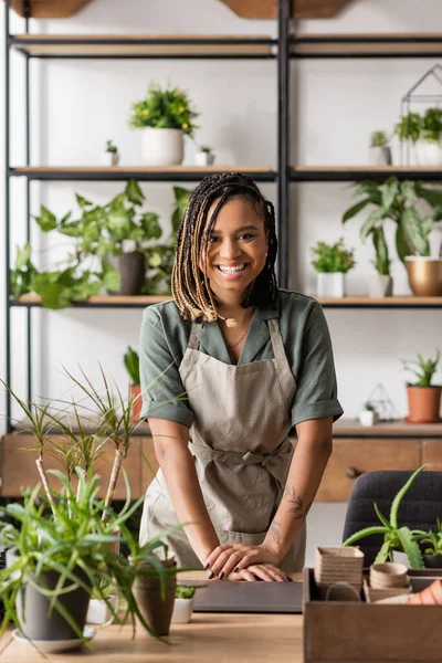 Happy african american florist in apron and dreadlocks smiling at camera near laptop in flower shop — Stock Photo