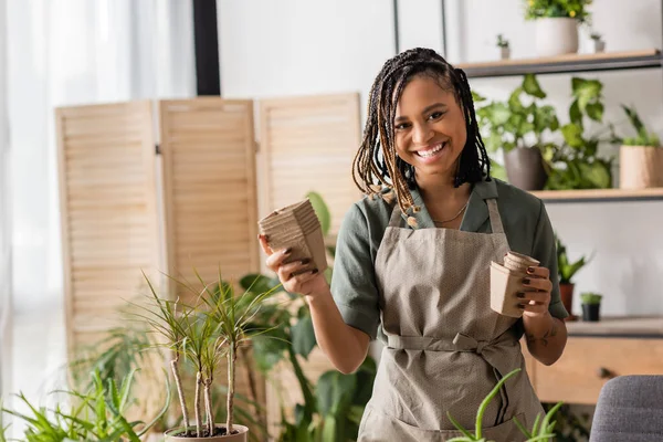 Cheerful african american florist with dreadlocks holding peat pots and looking at camera in flower shop — Stock Photo