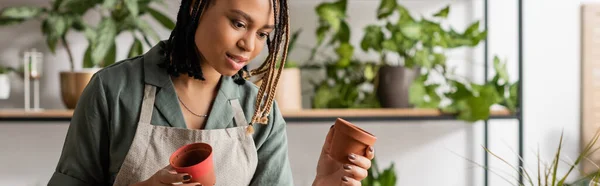 African american florist with braids choosing flowerpots while working in shop, banner — Stock Photo