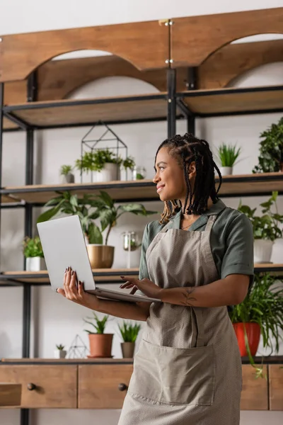 Happy african american florist in apron holding laptop and looking away in flower shop — Stock Photo