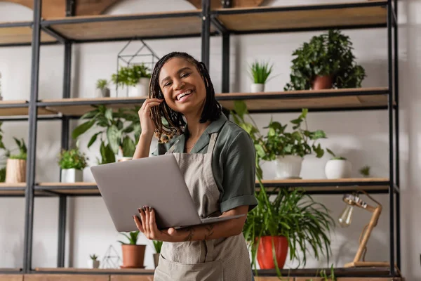 Cheerful african american florist with dreadlocks standing with laptop near rack with green potted plants on blurred background — Stock Photo