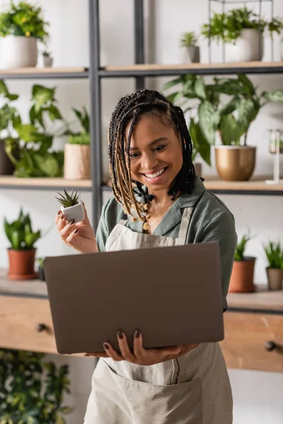 Fiorista afroamericano felice che tiene piccolo impianto in vaso durante videochiamata su computer portatile in negozio di fiori — Foto stock