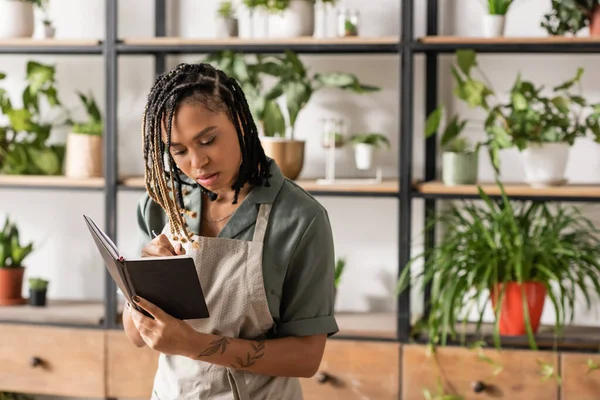 Giovane fiorista afroamericano in grembiule scrittura ordine nel taccuino in negozio di fiori — Foto stock